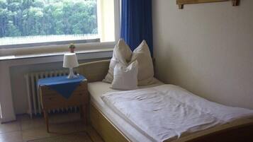 A small, neatly made bed with white linens, a bedside table with a lamp, and a window with a view of trees.