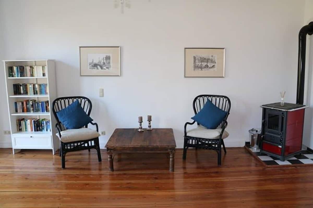 Quarto com piso de madeira, duas cadeiras pretas com almofadas azuis, uma pequena mesa de madeira, uma estante, duas fotos emolduradas na parede e um fogão vermelho e preto.