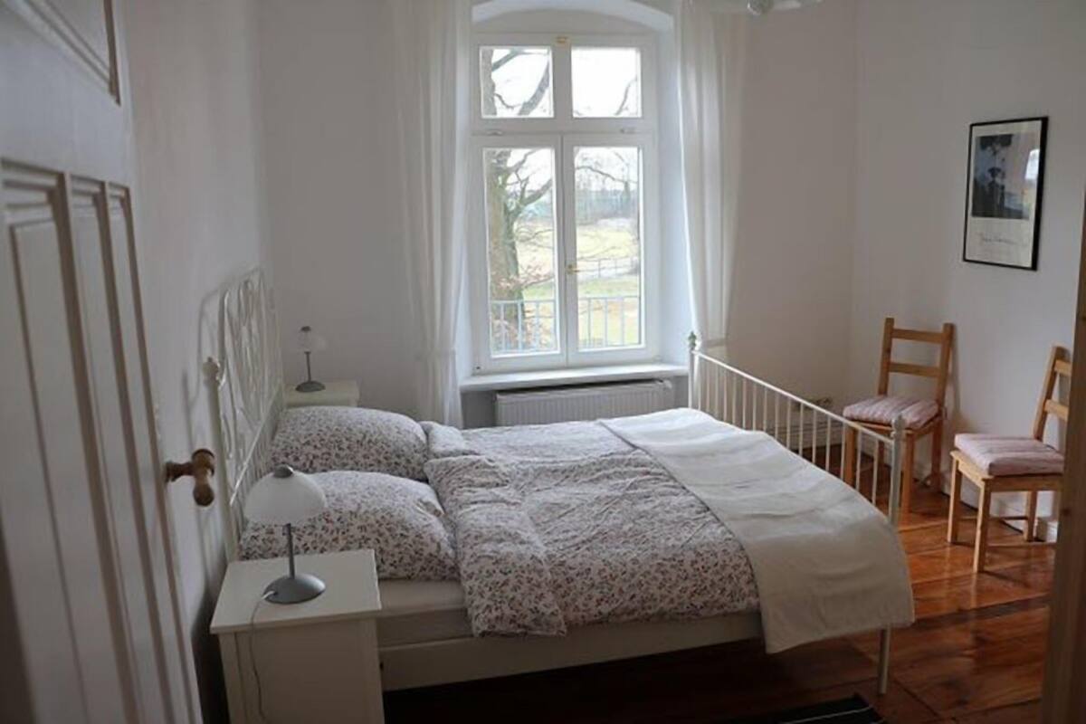 Quarto com cama, mesa de cabeceira, abajur, janela com cortinas e uma cadeira.