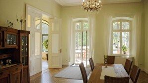 A dining area with a wooden table and chairs, a chandelier, and large windows with curtains.