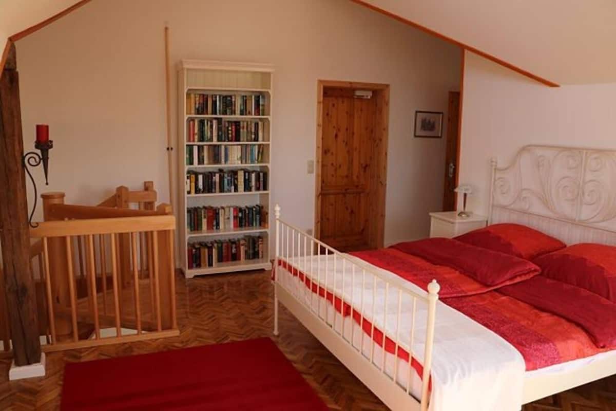 Quarto com berço de madeira, estante, porta de madeira e cama com roupa de cama vermelha.