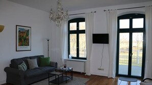 A living room with a dark sofa, a coffee table, a chandelier, a painting, and large windows with curtains.