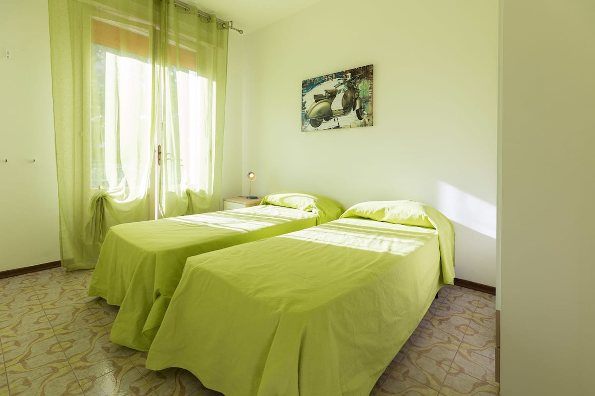 Una habitación con dos camas, una ventana con cortinas traslúcidas, un cuadro colgado en la pared y un suelo estampado.