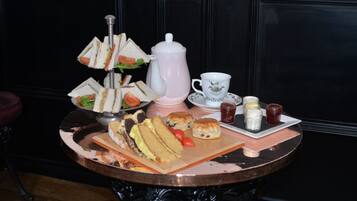 Una mesa con un soporte escalonado para sándwiches, una tetera, una taza, galletas y condimentos.