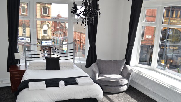 A modern hotel room with a large bed, a grey armchair, and a view of the city through large windows. - The Wrens (Leeds)