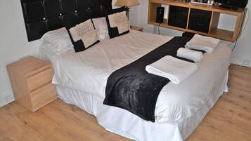 A neatly made bed with a black headboard, white bedding, and a black throw blanket. There is a wooden nightstand beside the bed and a wooden shelf with a television and other items.