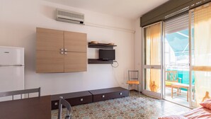 A compact room with a kitchenette, a bed, a chair, and a sliding glass door leading to an outdoor area.
