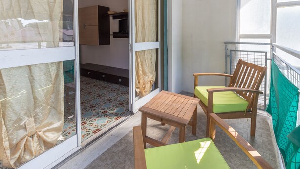 A balcony with a wooden table and two chairs, a tiled floor, and a view of the interior through a glass door.