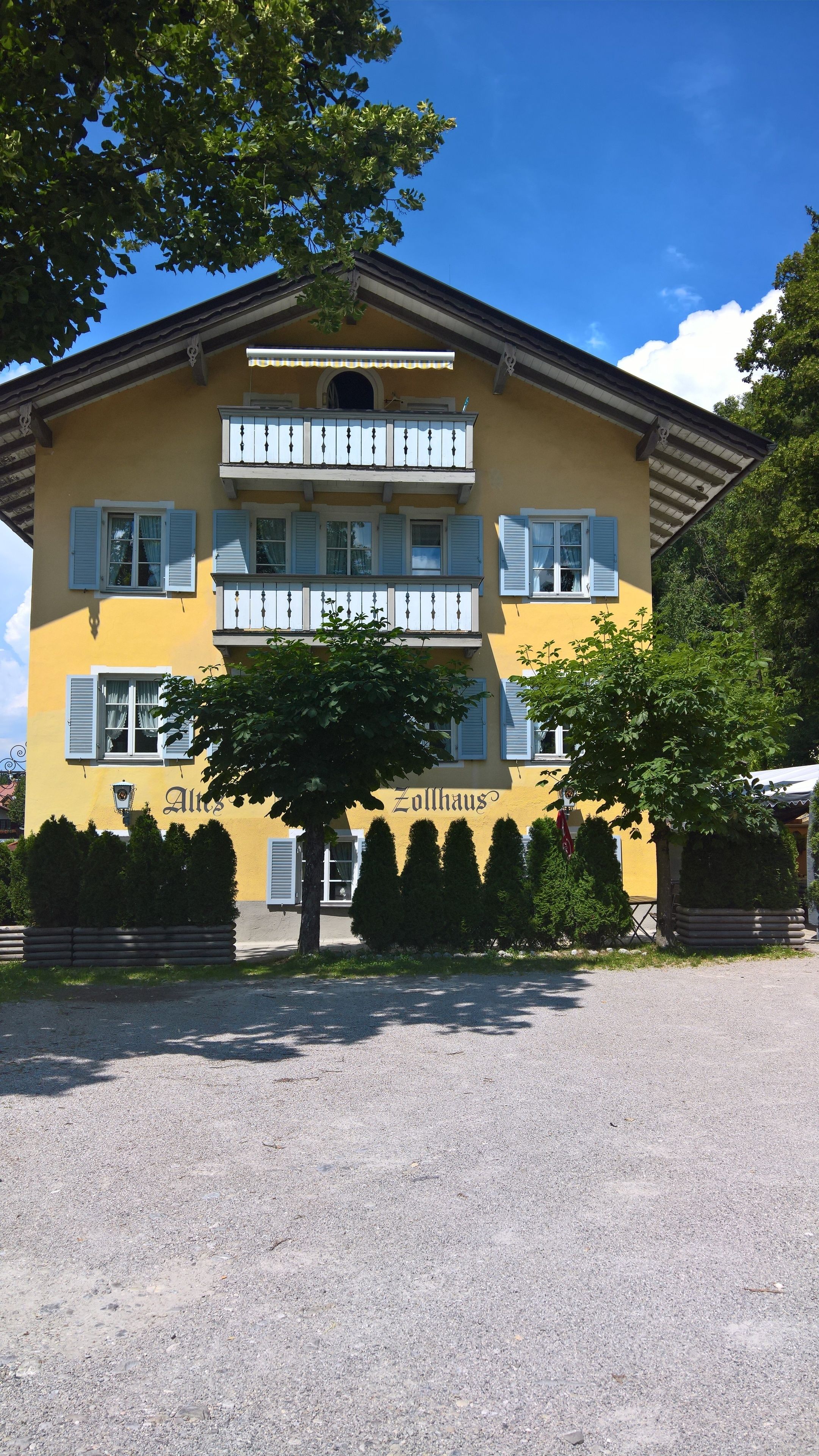 A yellow building with the name "Zollhaus" on the facade, featuring multiple windows with shutters, a balcony, and a gravel driveway.