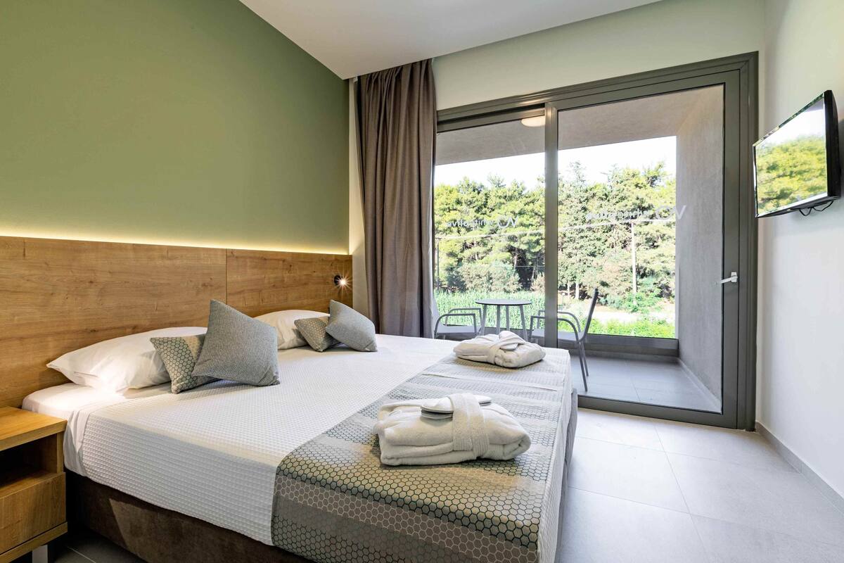 Habitación de hotel con una cama grande, cabecero de madera, balcón con mesa y sillas, y un televisor en la pared.