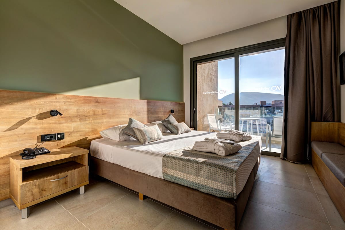Habitación de hotel moderna con una cama grande, una mesita de noche de madera con un teléfono, vistas a la ciudad a través de amplias ventanas y un balcón.