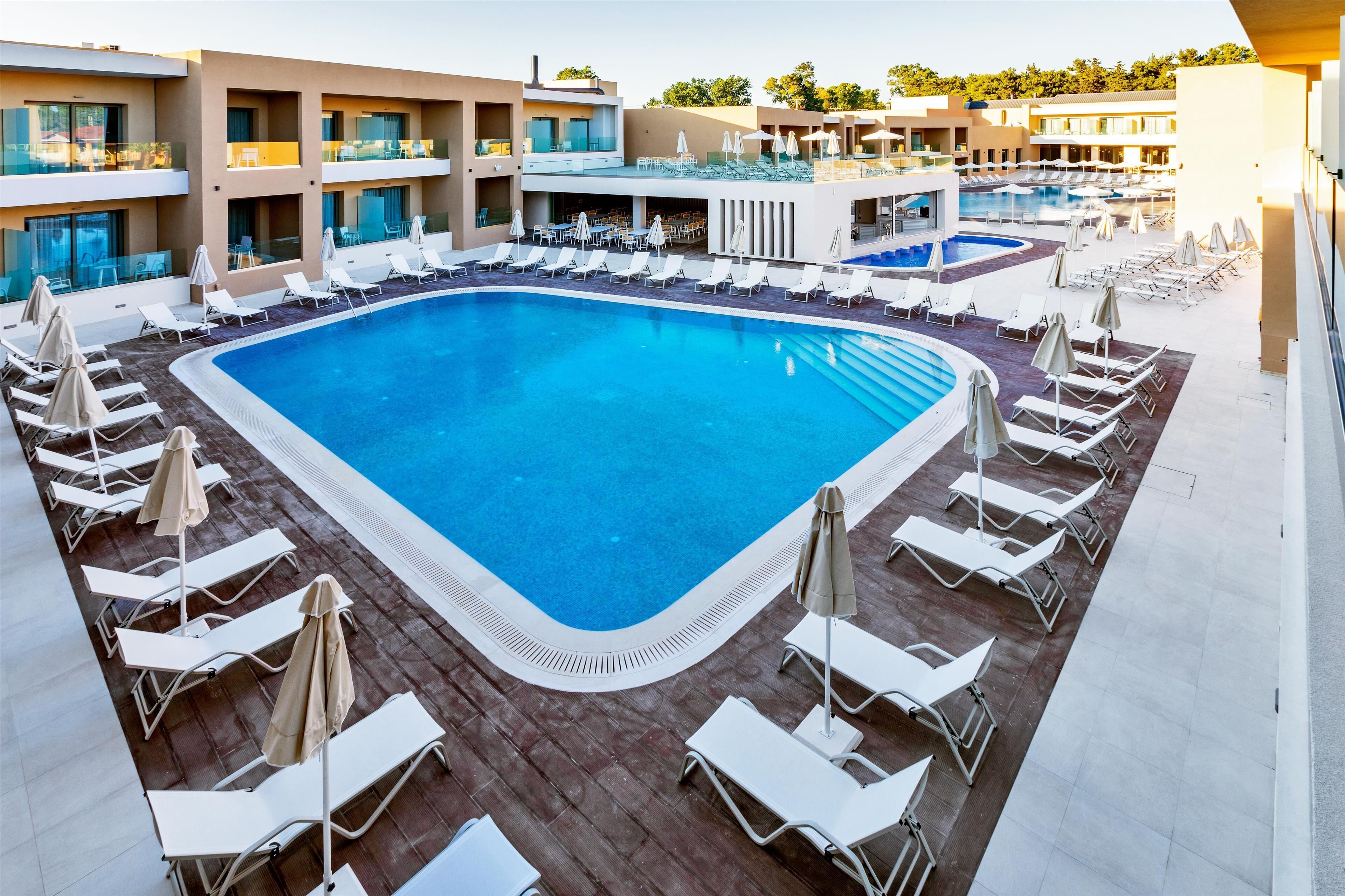 A large rectangular swimming pool surrounded by white sun loungers and umbrellas, with a multi-story building in the background.