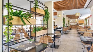 A spacious restaurant with high tables and wicker chairs, wooden beams on the ceiling, and potted plants on shelves.