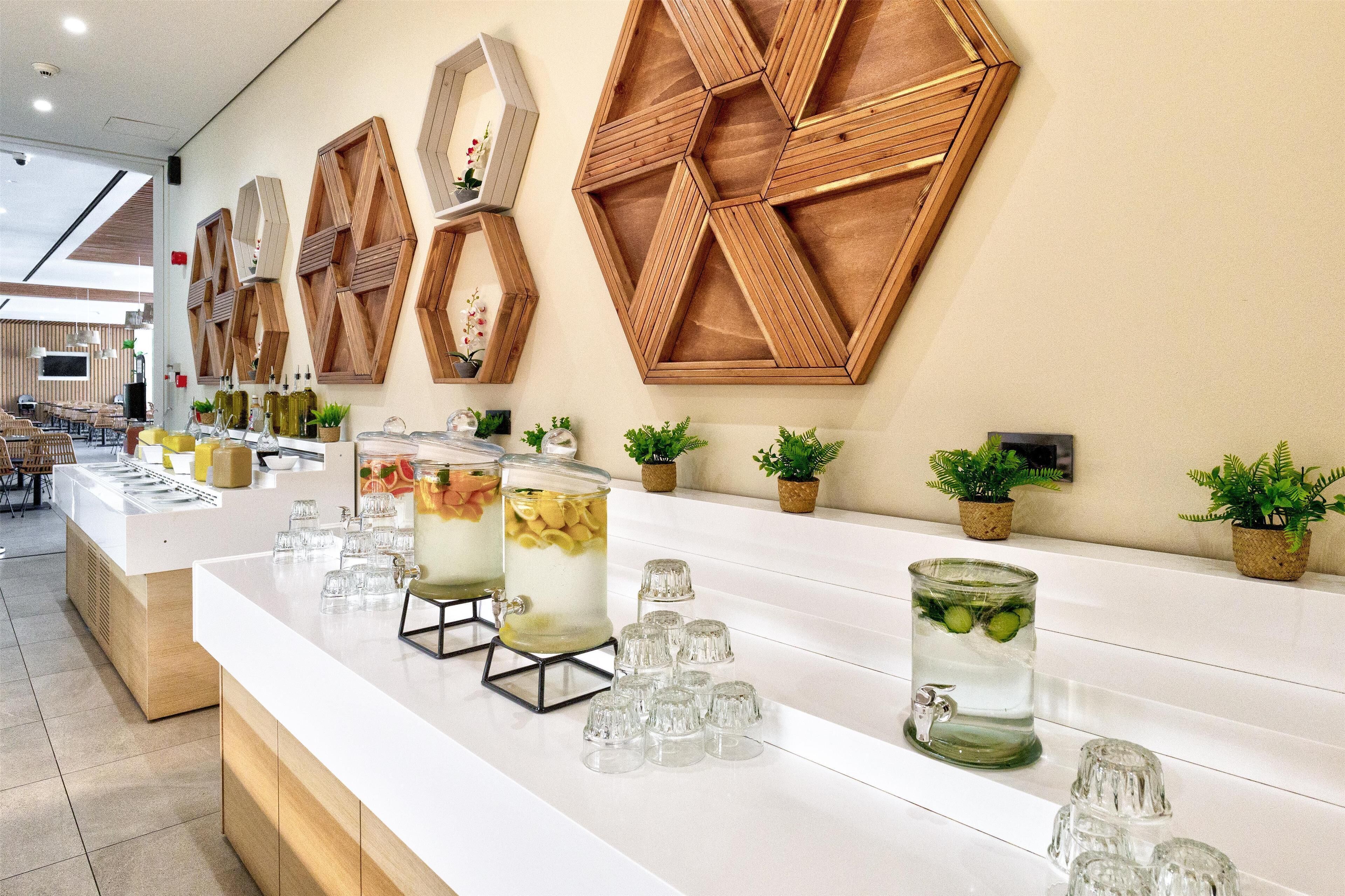 Uma área de bar moderna, com decoração hexagonal em madeira na parede, balcão branco, diversos copos e vasos com plantas.