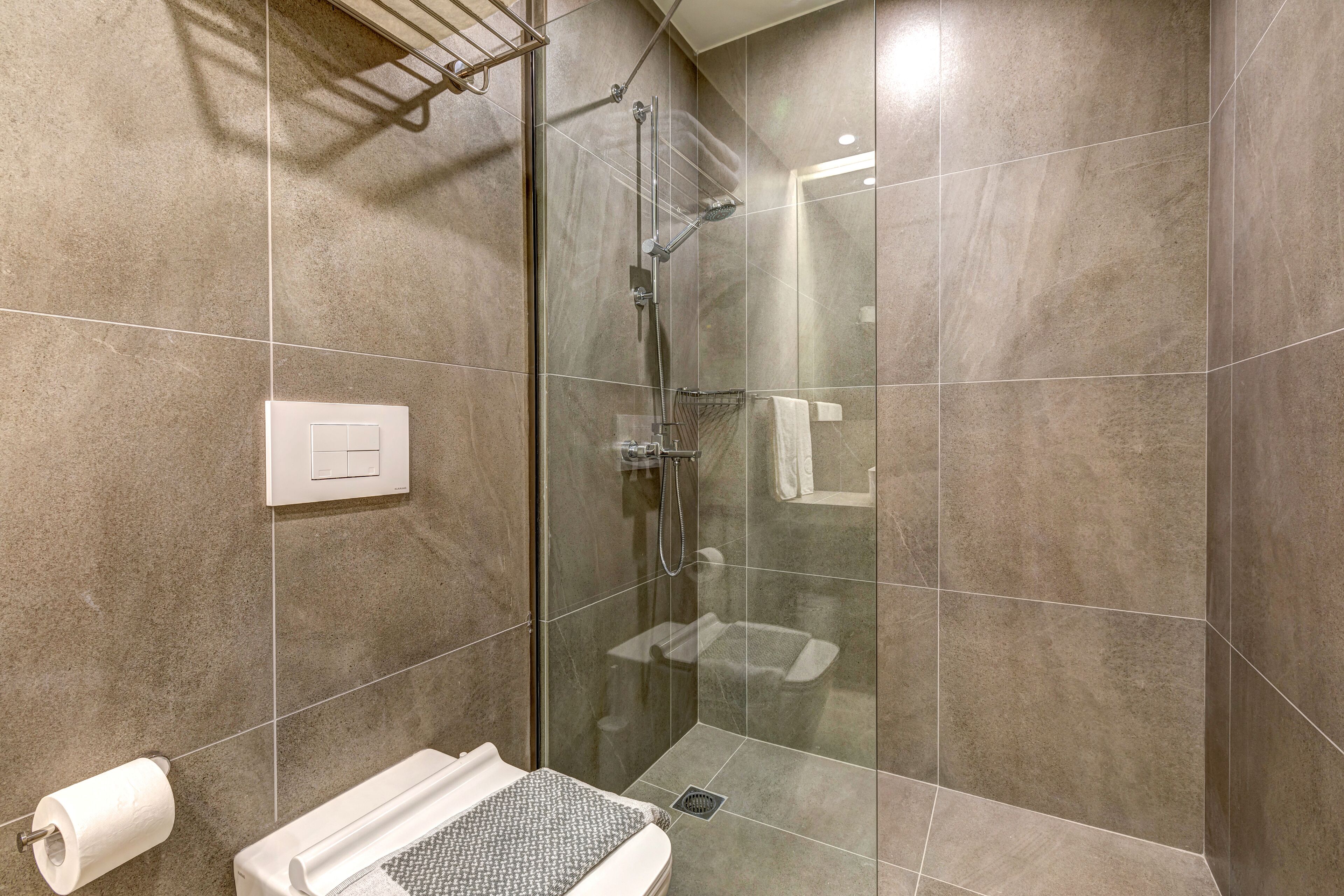 A modern bathroom with a glass-enclosed shower, a toilet with a towel rack, and a wall-mounted light switch.