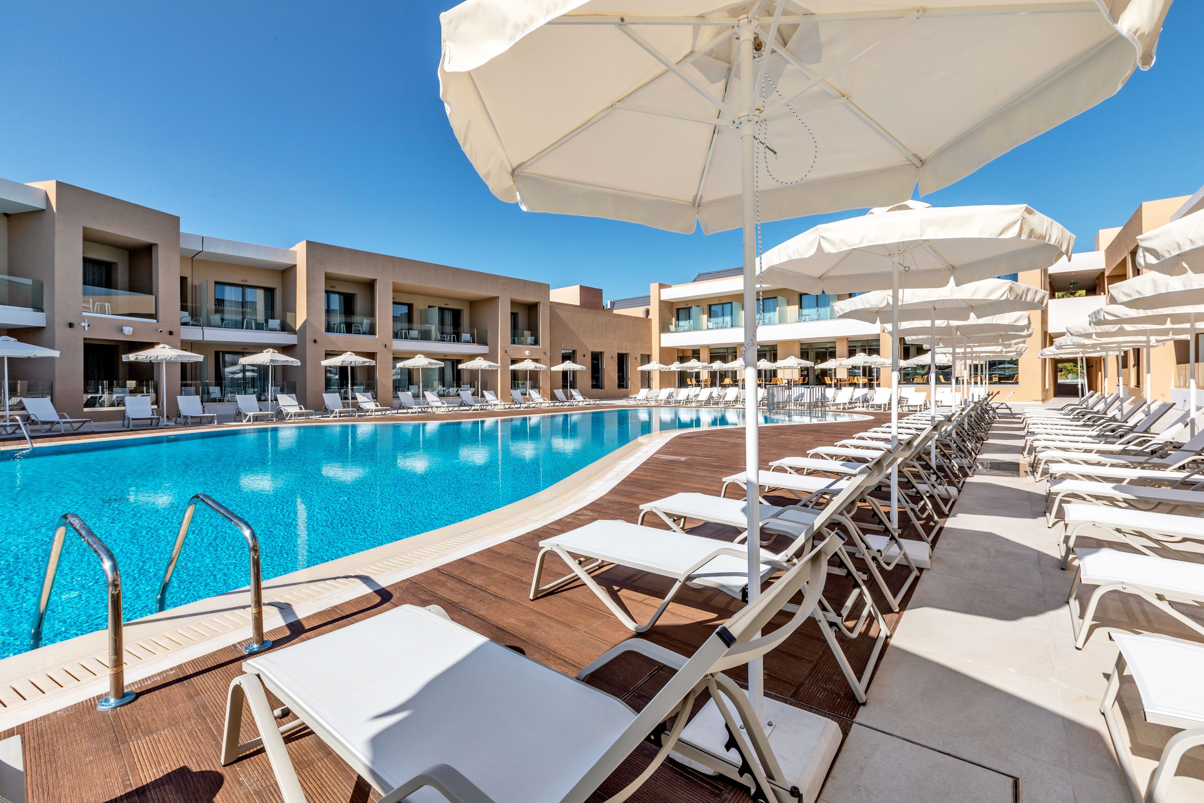 A poolside area with sun loungers and umbrellas, a clear blue pool, and a modern building with balconies.
