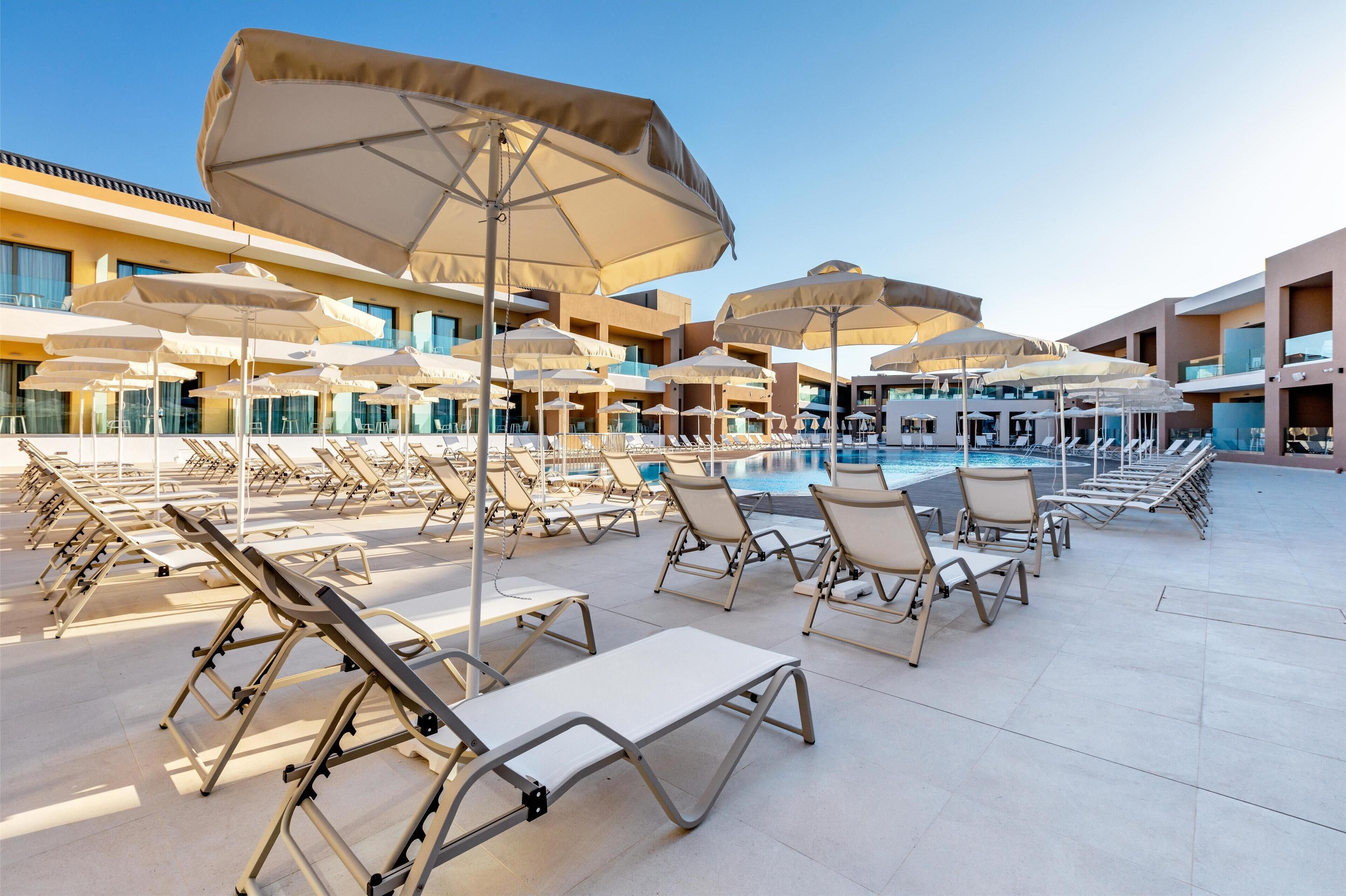 Uma área na piscina com espreguiçadeiras e guarda-sóis, céu azul límpido e um edifício moderno ao fundo.
