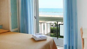 Una habitación de hotel con cama, balcón con cortinas azules y vista a la playa.