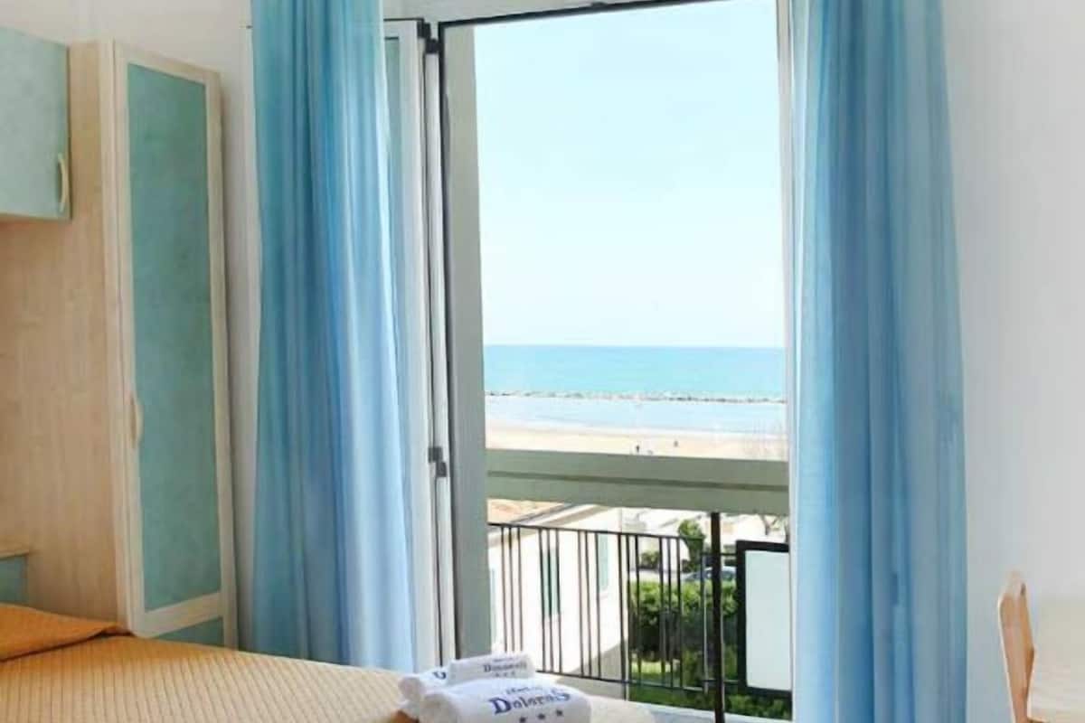 A hotel room with a bed, a balcony with blue curtains, and a view of the beach.