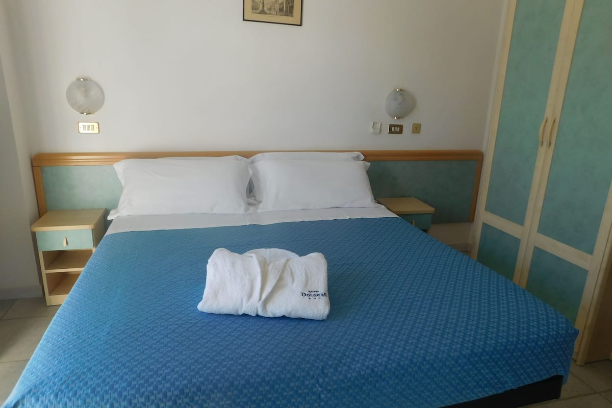 A neatly made bed with a blue bedspread and two white pillows, a bedside table with a lamp, and a wardrobe with a green door.