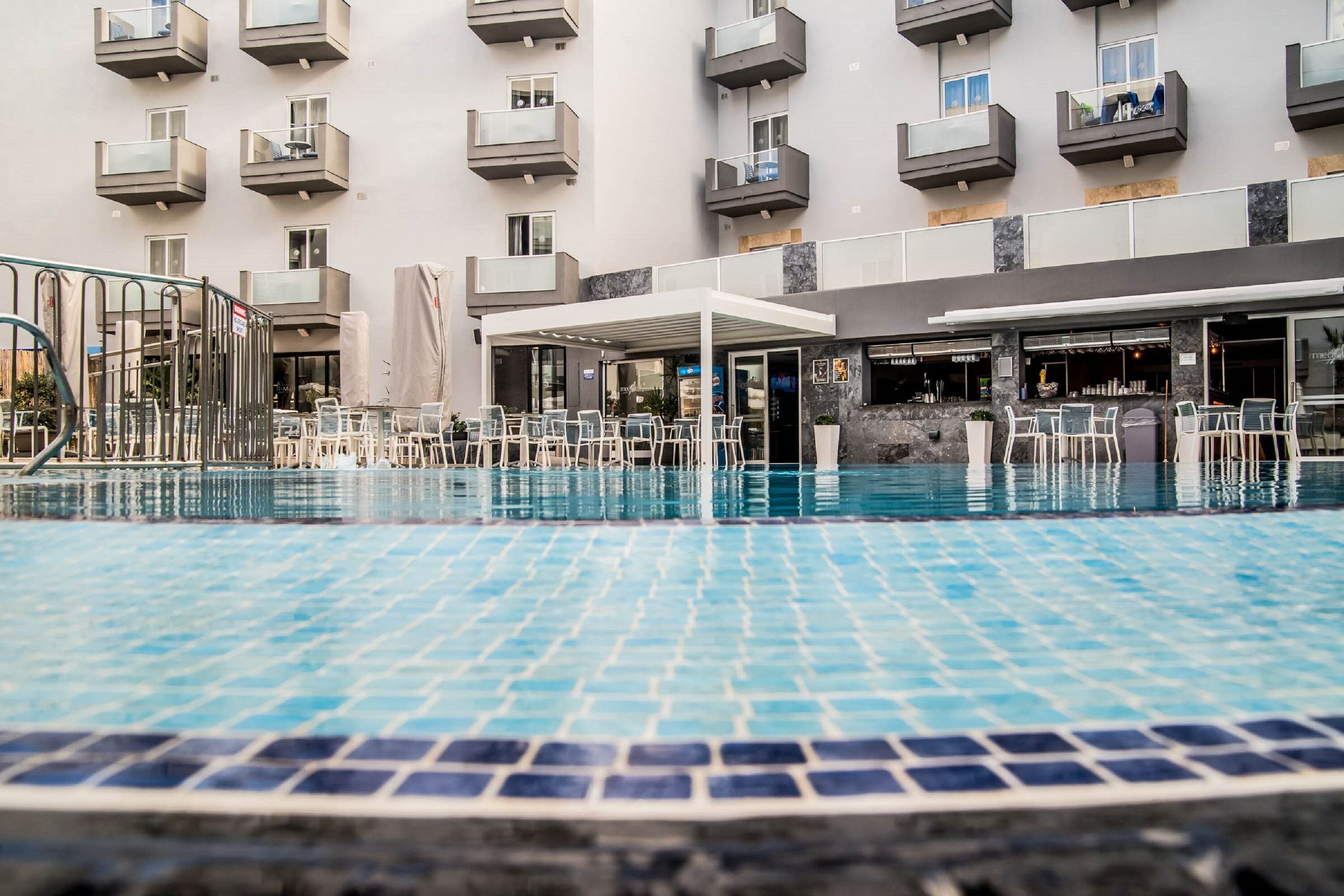 Un'area piscina con pavimento piastrellato, arredi esterni e un edificio con balconi e un bar.