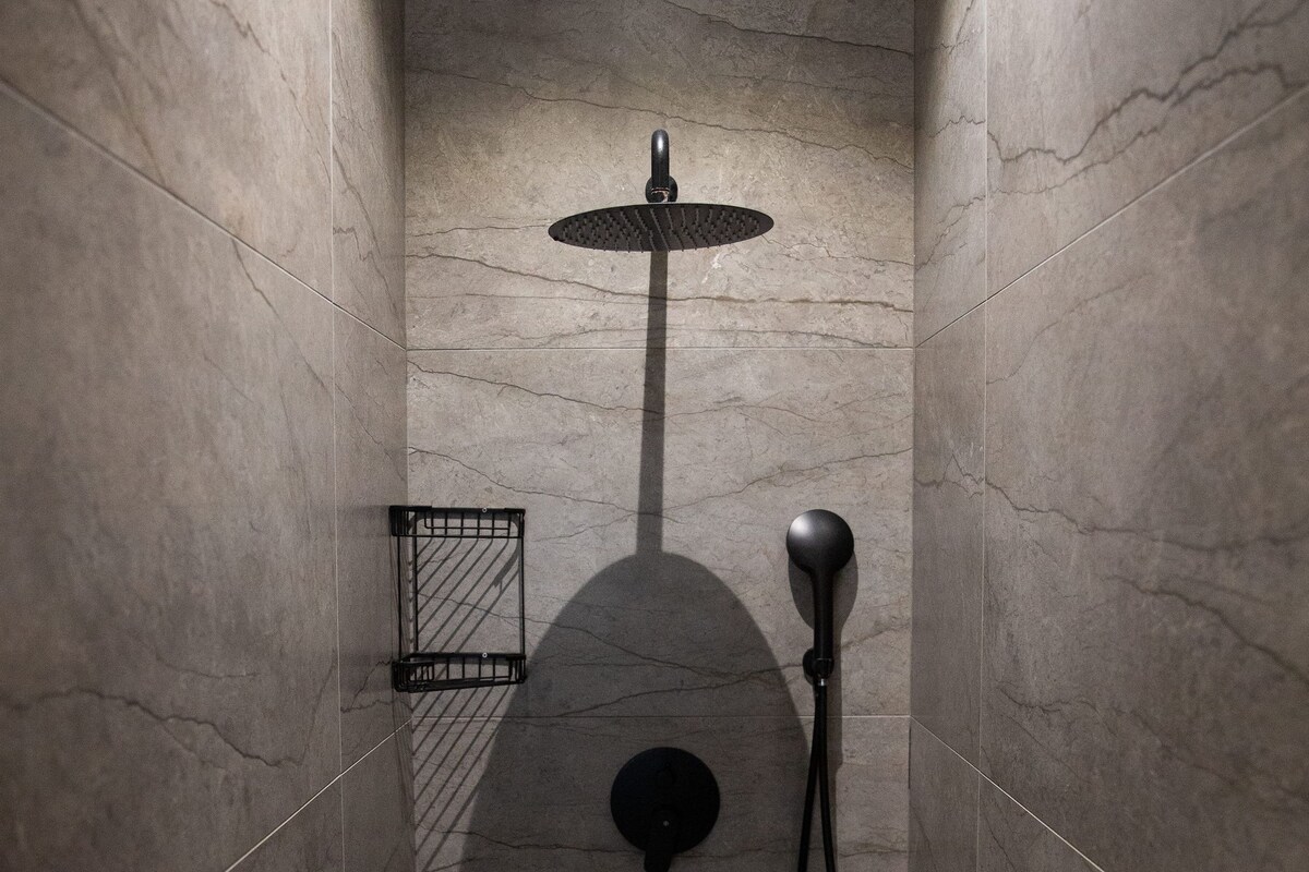 A shower with a handheld showerhead, a wall-mounted faucet, and a small shelf.