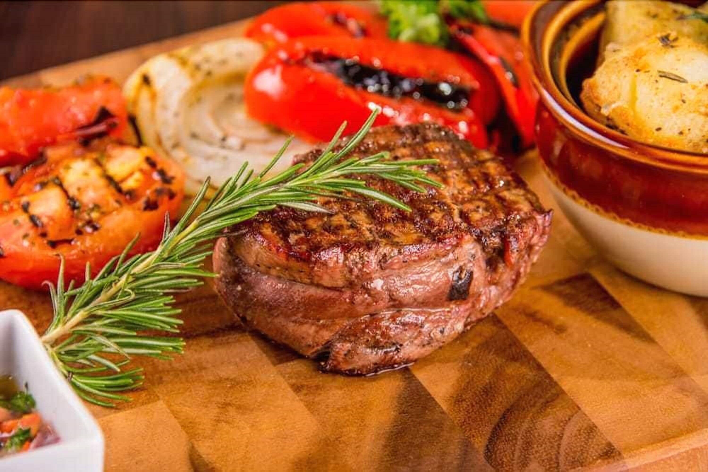 A steak with a sprig of rosemary on a wooden board, accompanied by grilled vegetables and a baked potato.
