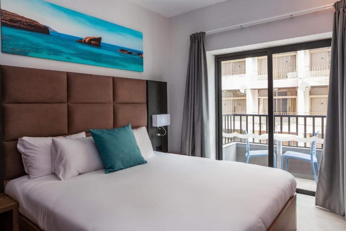 Habitación de hotel con una cama grande, un cuadro de una escena costera, un balcón con sillas y vistas a un edificio.