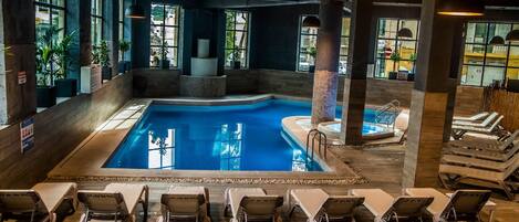 An indoor swimming pool with lounge chairs and a hot tub, surrounded by large windows and potted plants.