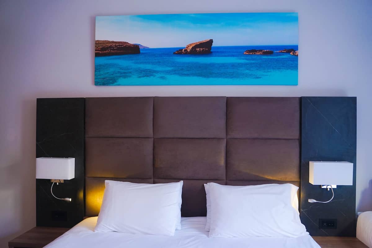 Ett hotellrum med en stor säng, två sänglampor, en sänggavel i marmor och en tavla som föreställer ett kustlandskap.