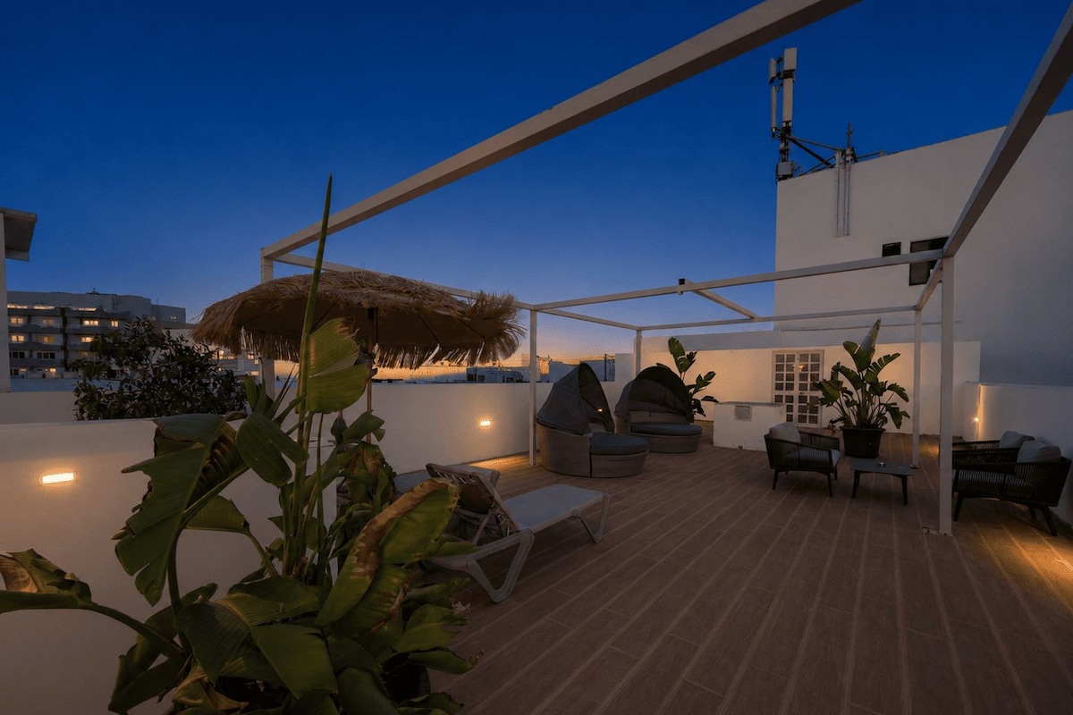 A rooftop patio with a thatched umbrella, outdoor seating, and a view of the cityscape.