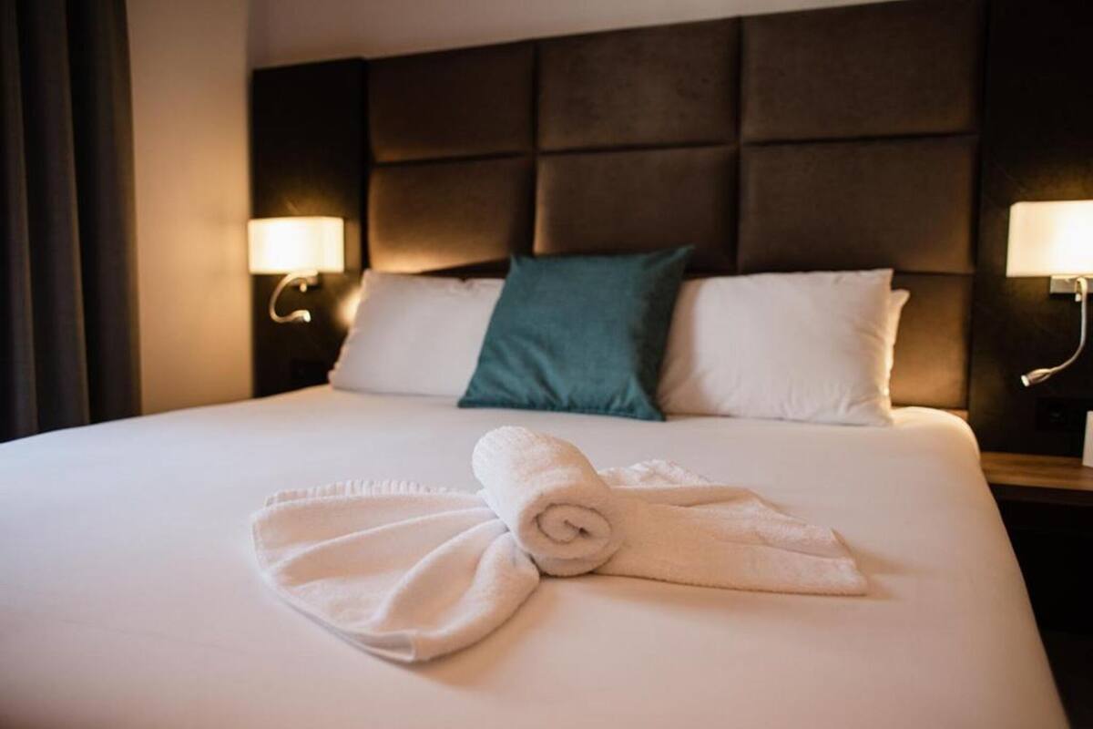 Habitación de hotel con una cama grande, dos almohadas, una toalla enrollada y dos lámparas fijadas a la pared.