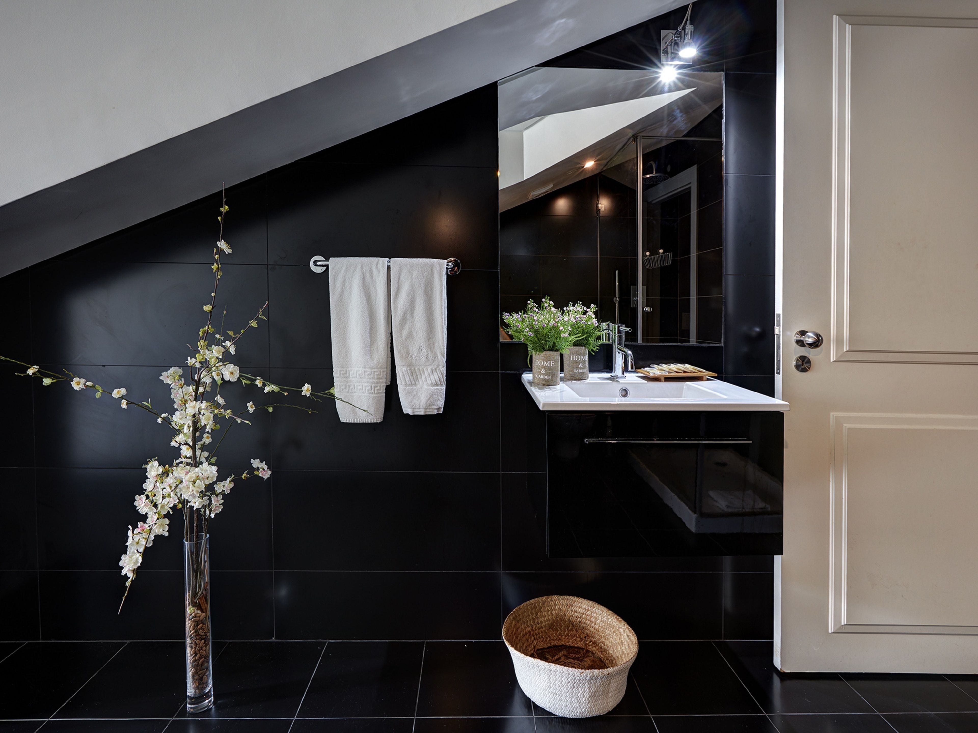 A modern bathroom with black tiles, a white sink, a towel rack, a mirror, and a vase with flowers.