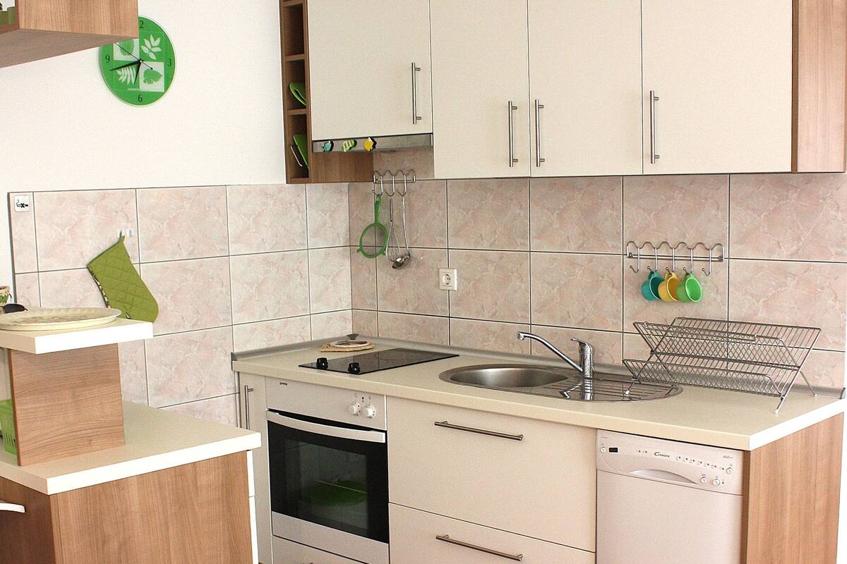 Une cuisine avec des armoires blanches, un évier, une cuisinière et un lave-vaisselle. Il y a une horloge verte accrochée au mur et une chaise près de la table.
