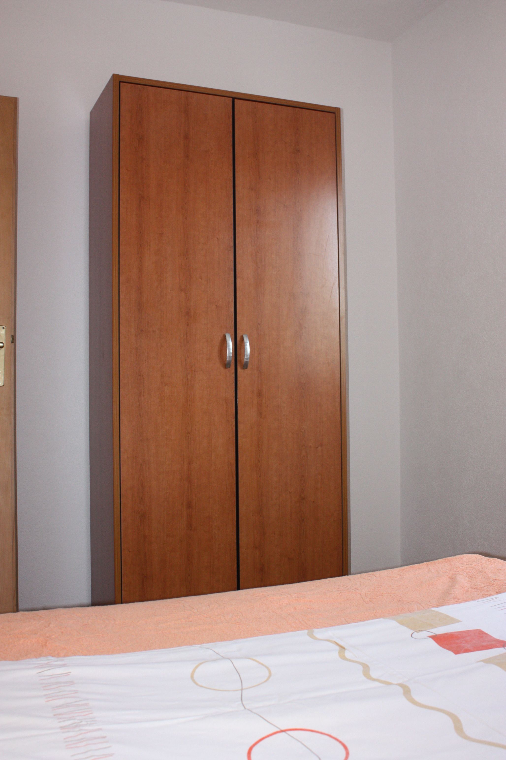 A bedroom with a wooden wardrobe, a bed with a patterned bedspread, and a plain wall.
