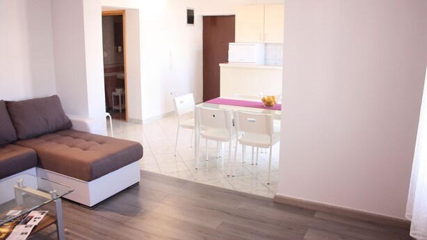 A modern living room with a sectional sofa, a glass coffee table, and a dining area with a pink tablecloth and white chairs. - Apartments Miki Pag (Pag)