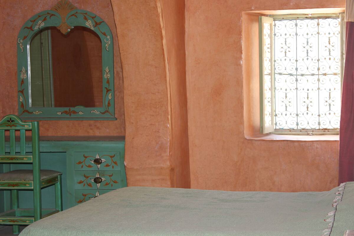 A room with a bed, a dresser, a chair, a mirror, and a window with a metal screen.