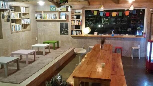 Un café acogedor con mesas y sillas de madera, una pizarra con escritura y estantes llenos de libros.