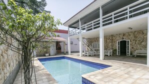 A swimming pool with a stone wall and a tree on one side, and a covered patio area with a staircase. - Alameda dos Eucalipitos (Rio de Janeiro)