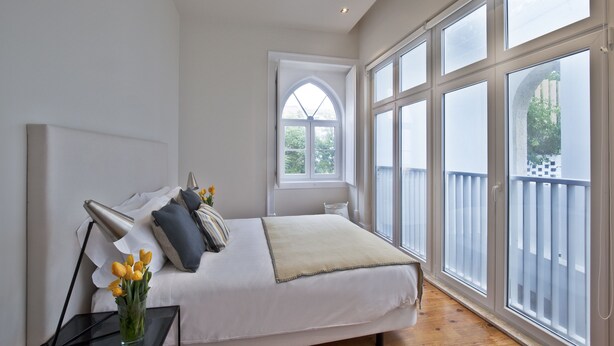 A bedroom with a large bed, a bedside table with a lamp, a window with a view of greenery, and a balcony with a railing.