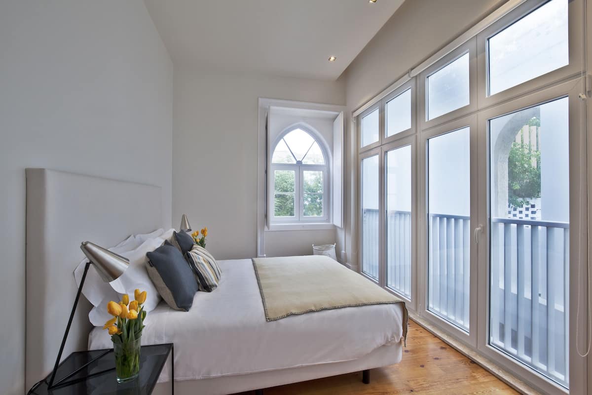 A bedroom with a large bed, a bedside table with a lamp, a window with a view of greenery, and a balcony with a railing.