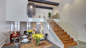 A spacious dining area with a wooden table, chairs, and a hanging light fixture. A staircase with glass railings leads to an upper level.