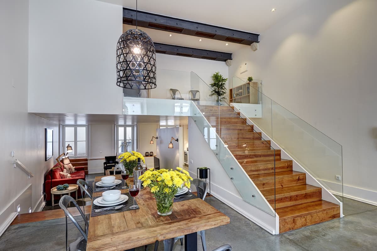 A spacious dining area with a wooden table, chairs, and a hanging light fixture. A staircase with glass railings leads to an upper level.