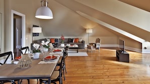 A spacious loft with a high ceiling, wooden flooring, a dining area with a long table, and a cozy living space with a sofa and TV.