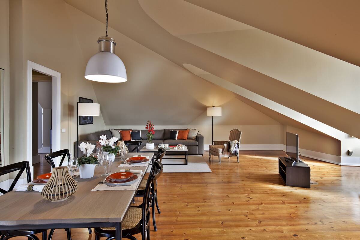 A spacious loft with a high ceiling, wooden flooring, a dining area with a long table, and a cozy living space with a sofa and TV.