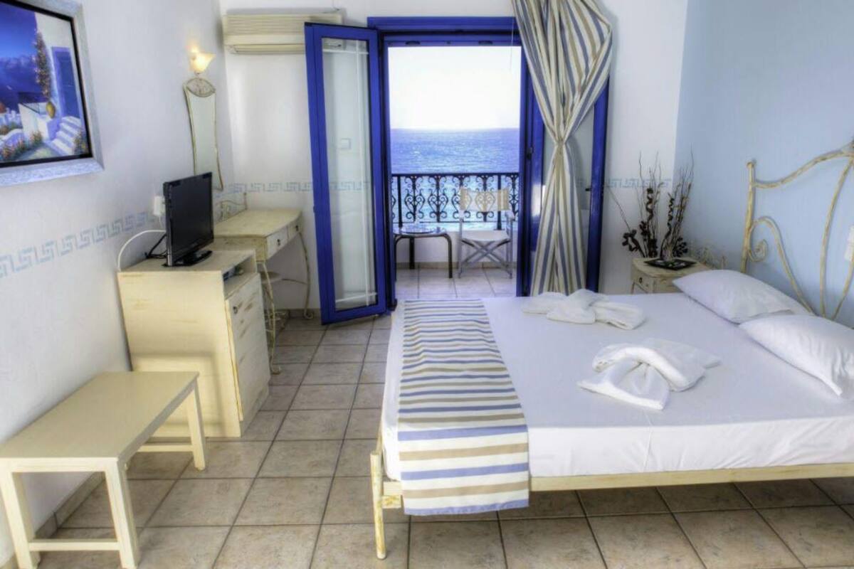 A hotel room with a bed, a desk, a chair, a TV, and a view of the sea through an open door.
