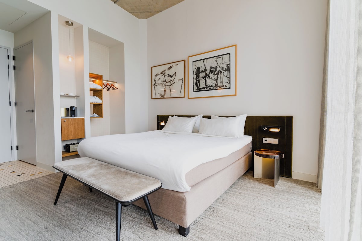 Moderni hotellihuone, jossa on suuri sänky, yöpöydät, penkki ja taideteoksia seinillä.