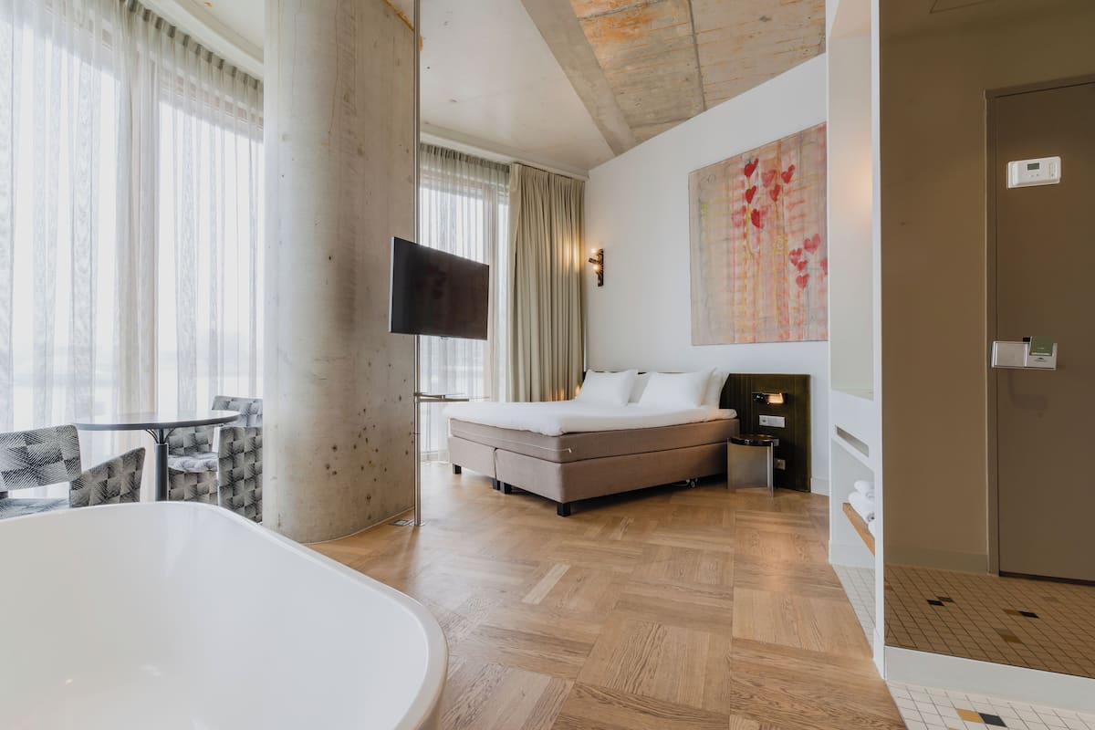 Moderni hotellihuone, jossa on suuri sänky, taulutelevisio, kylpyamme ja ruokailutila tuoleineen.