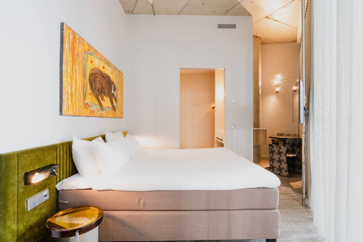 Hotellihuone, jossa on sänky, sängynpääty, seinään kiinnitetty valaisin, taulu ja avoimesta ovesta näkyvä kylpyhuone.