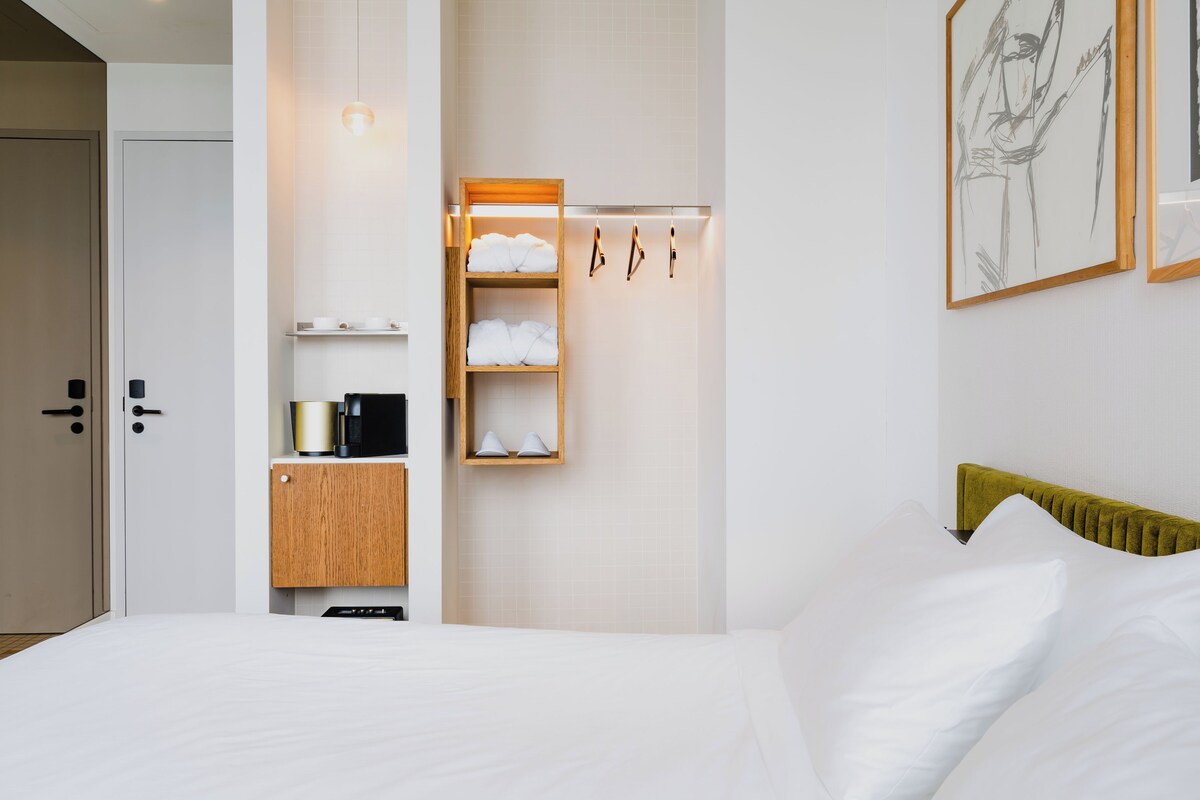 Quarto de hotel com cama, guarda-roupa de madeira, prateleira com toalhas, máquina de café e luminária fixada na parede.
